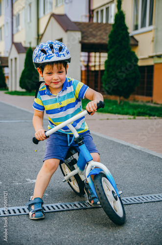 Fototapeta Naklejka Na Ścianę i Meble -  Cute boy of three years in a protective helmet rides a runbike (bicycle) in the courtyard of a multi-storey building. Children's summer street games.