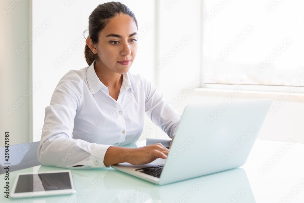 Serious female office employee working on computer. Young Latin businesswoman sitting at workplace, using laptop, typing, looking at screen. White office interior concept Serious female office employee working on computer. Young Latin businesswoman sitting at workplace, using laptop, typing, looking at screen. White office interior concept