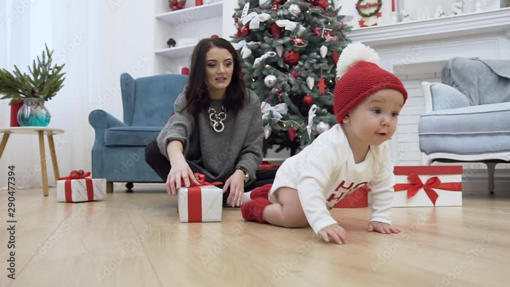Good-looking mother sitting on the floor near christmas tree with her adorable infant which trying to catch red ball
