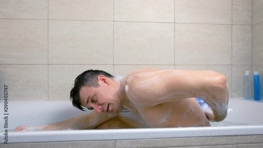Man is washing his back washcloth with shower gel sitting in bathtub. side view.