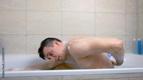 Man is washing his back washcloth with shower gel sitting in bathtub. side view.