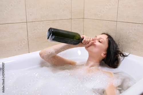 Funny young adult woman drinking wine from bottle lying relaxing in bath tub with foam. Smiling tipsy person in depression at home in evening. Concept of disappointment in life. Midlife crisis.