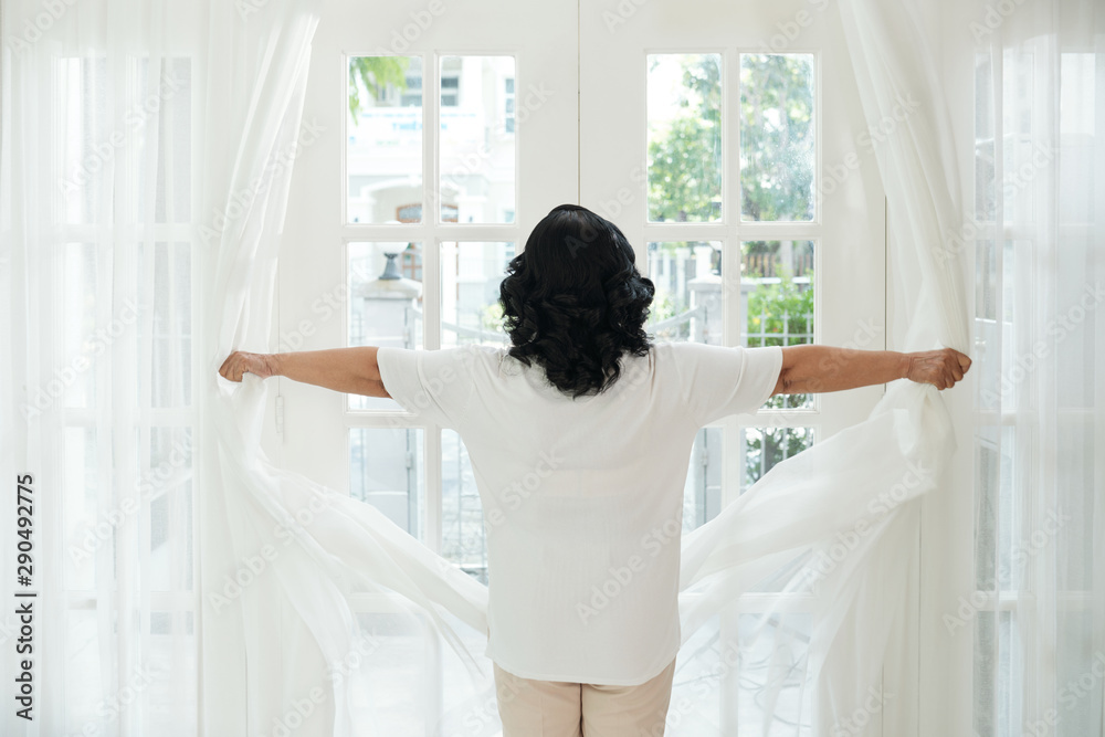 Rear view of woman with short dark hair opening curtains and looking through the window in her bedroom at home Rear view of woman with short dark hair opening curtains and looking through the window in her bedroom at home
