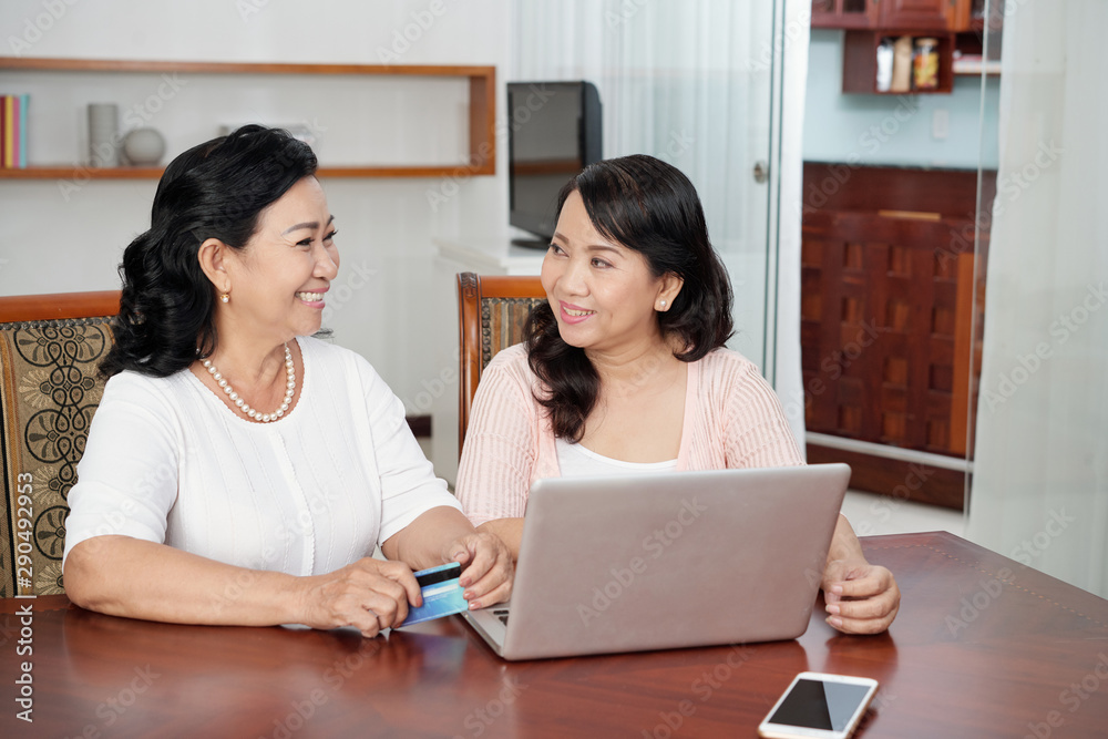 Two Asian senior women talking to each other while sitting at the table with laptop they are going to pay online by credit card Two Asian senior women talking to each other while sitting at the table with laptop they are going to pay online by credit card