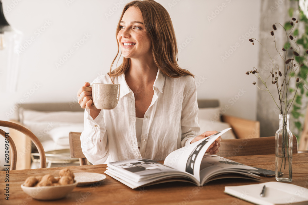 Woman sit indoors in office reading magazine drinking coffee.