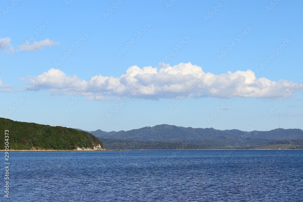 Green cape in blue sea under the clear blue sky with one wide cloud above. Sunny day, calm blue water. Background, wallpaper , template. Green cape in blue sea under the clear blue sky with one wide cloud above. Sunny day, calm blue water. Background, wallpaper , template.