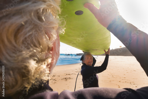 couple of seniors ready to go to surf at the beach holding together a big green surf table - trying their firts time in a surf lessons in their vacation photo