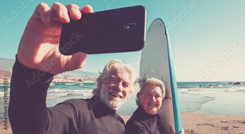 happy couple of seniors at the beach trying to go surf and having fun together - mature woman and man married taking a selfie with the wetsuits and surftables with sea or ocean at the background photo