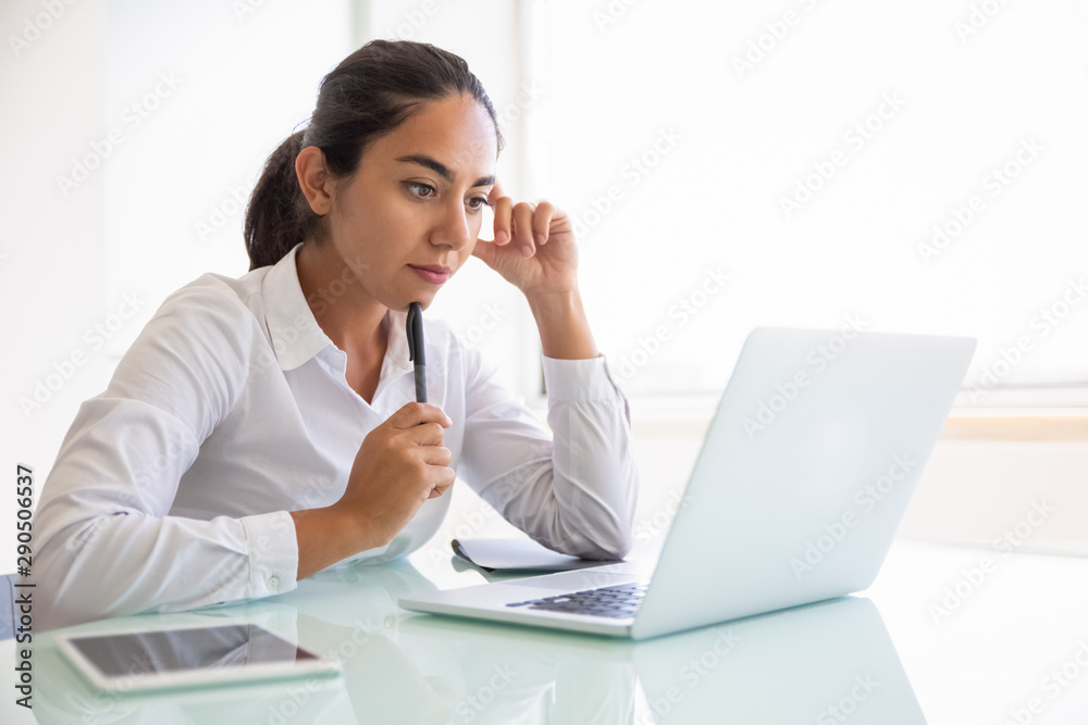 Serious pensive businesswoman watching content on computer. Young Latin business woman sitting at workplace, using laptop, looking at screen. Digital communication concept Serious pensive businesswoman watching content on computer. Young Latin business woman sitting at workplace, using laptop, looking at screen. Digital communication concept