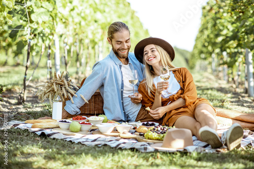 Beautiful couple having romantic breakfast with lots of tasty food and wine, sitting together on the picnic blanket at the vineyard on a sunny morning