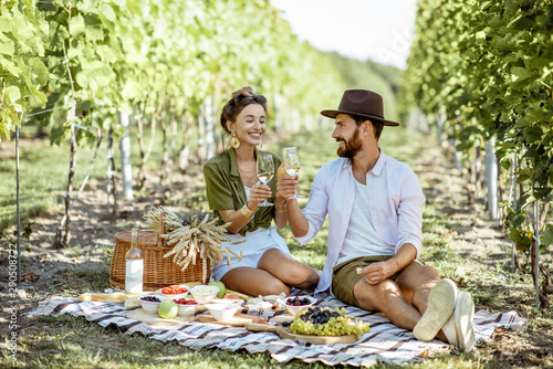 Beautiful couple having romantic breakfast with lots of tasty food and wine, sitting together on the picnic blanket at the vineyard on a sunny morning