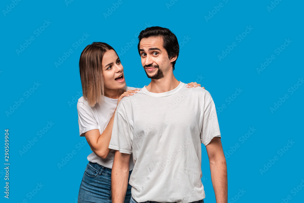 Young blond woman tells somthing important to her brunet bearded boyfriend with mustaches in white t-shirts and blue jeans isolated over blue background. Concept of perfect couple. Young blond woman tells somthing important to her brunet bearded boyfriend with mustaches in white t-shirts and blue jeans isolated over blue background. Concept of perfect couple.