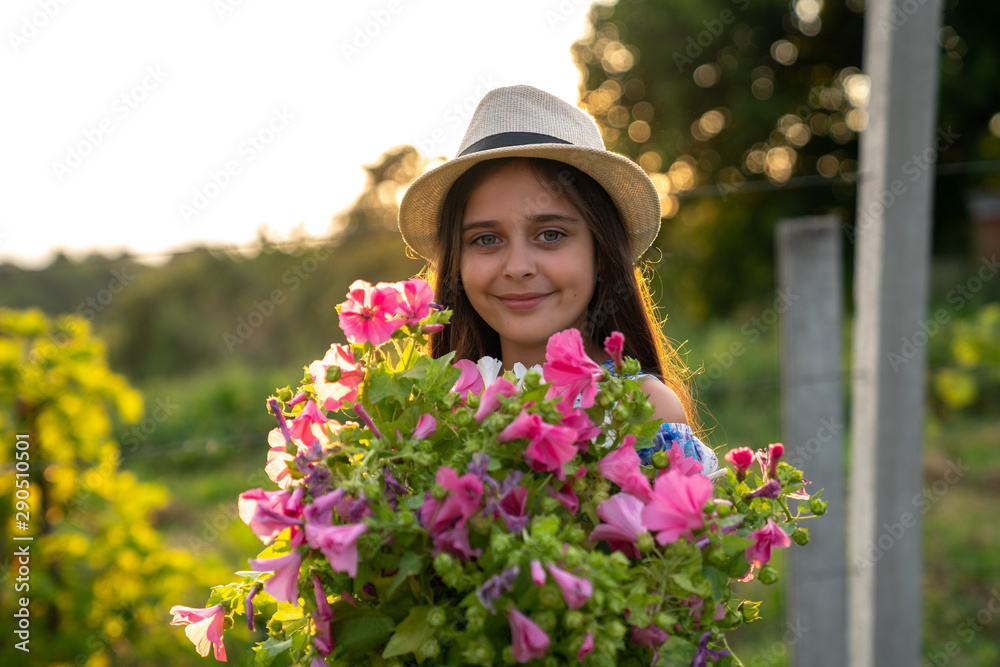 Half length portrait of smiling cute little girl with healthy skin in white hat and dress that holding a large bouquet of pink flowers, landscape on the background