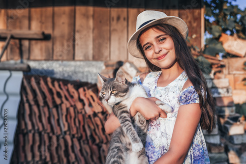 Happy smiling young girl in white hat and dress with flowers print holding a white and grey cat on her hands, looking at the camera, wooden wall and old bricks on the background