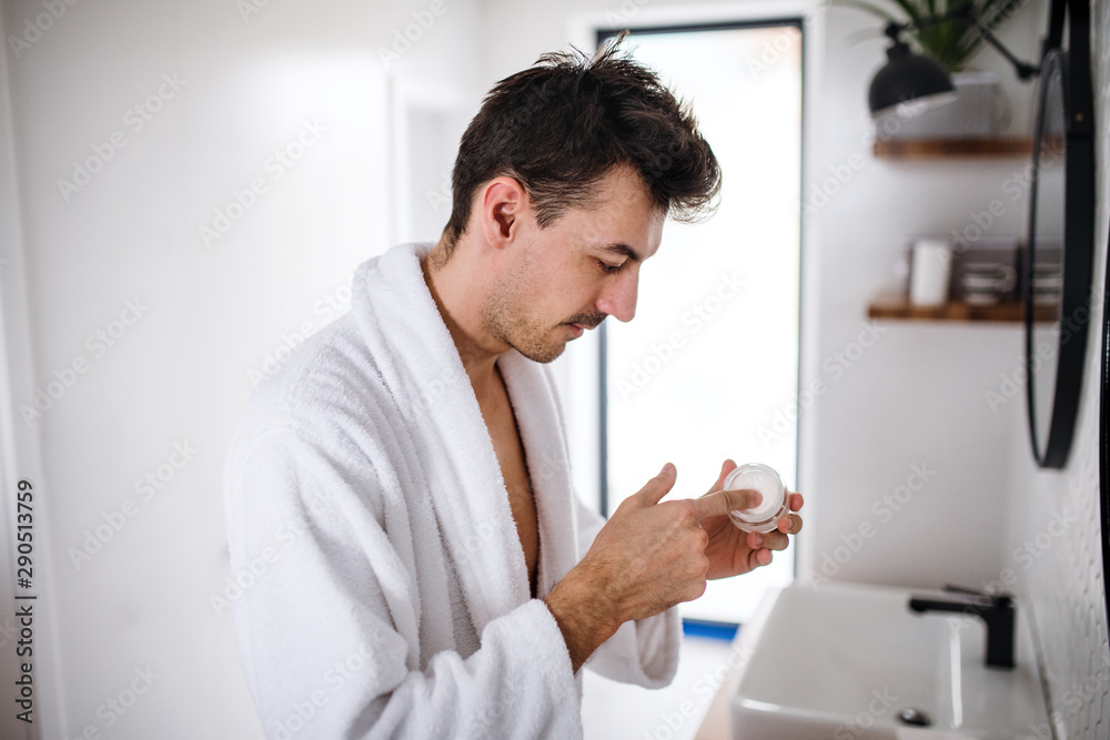 Young man putting cream on face in the bathroom in the morning, daily routine.