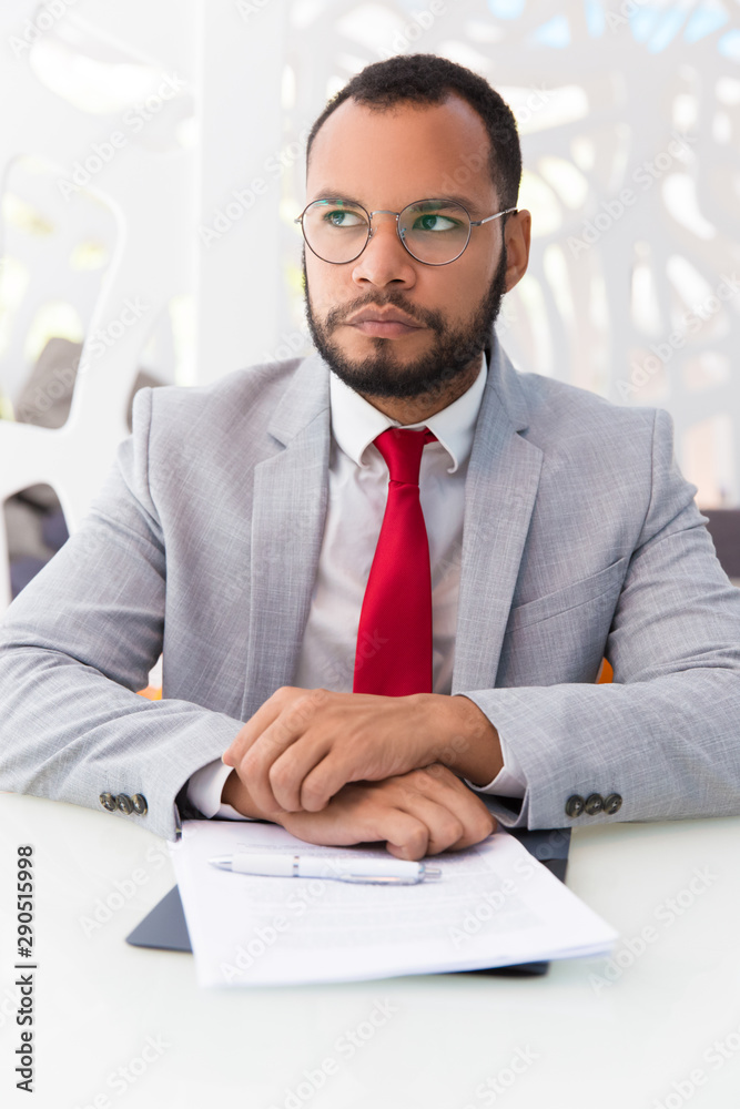 Pensive businessman thinking over agreement. Business man wearing suit and eyeglasses, sitting at office table with papers and pen and looking up. Business decision concept Pensive businessman thinking over agreement. Business man wearing suit and eyeglasses, sitting at office table with papers and pen and looking up. Business decision concept