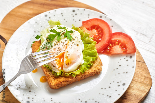 Poached egg, benedict on toasted white bread toast with salad and spices on a plate on a light wooden background.