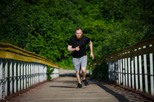 active young adult caucasian man with beard wearing black shirt and sneakers running along wooden bridge in natural park during cardio sport training