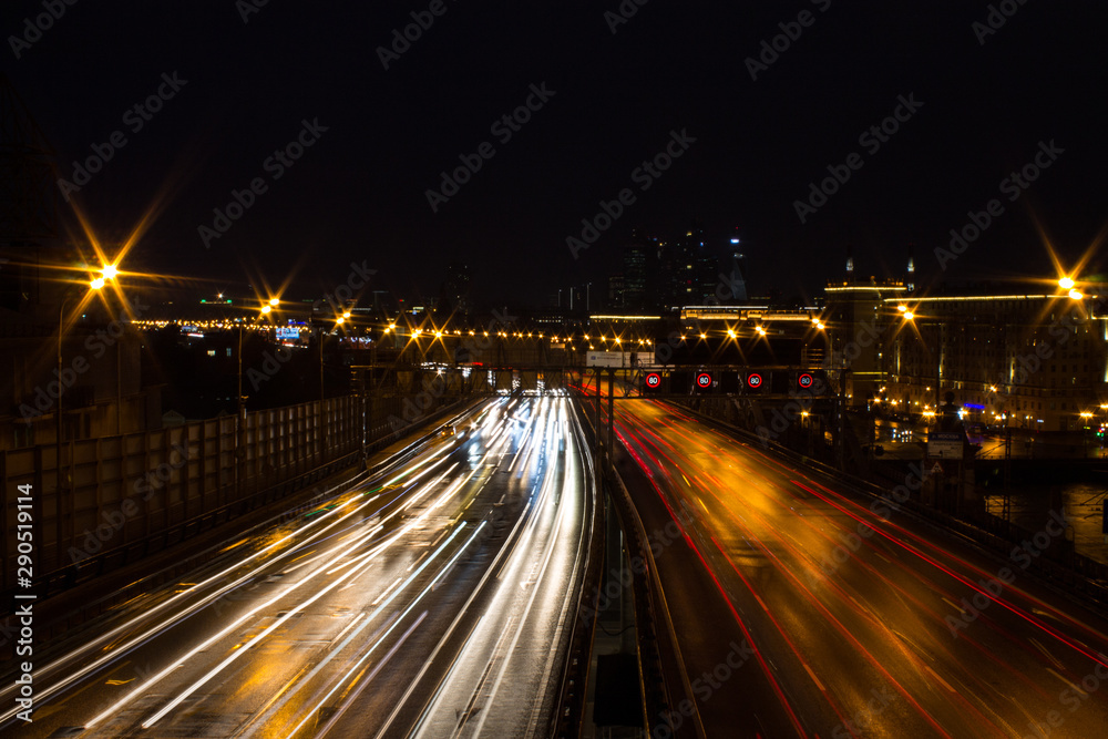 Panoramic view on a city highway with glowing lines of light from headlamps and architecture against the night dark sky