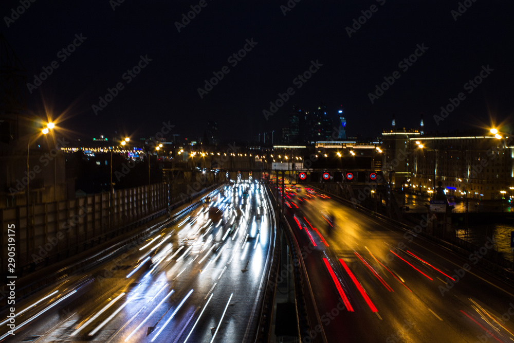 Fototapeta premium Panoramic view on a city highway with glowing lines of light from headlamps and architecture against the night dark sky