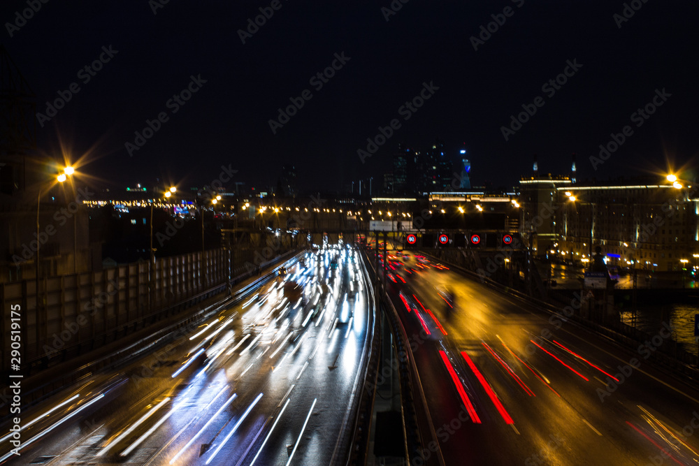Panoramic view on a city highway with glowing lines of light from headlamps and architecture against the night dark sky