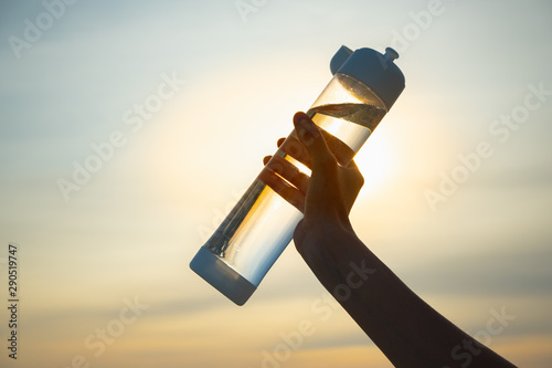 Human hand holds a water bottle against the setting sun. Close up of a reusable water bottle in a human hand, concept of thirst, rehydration and decreasing single use plastic