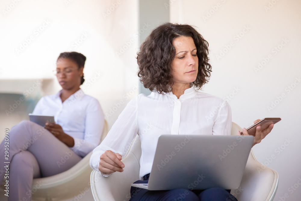 Focused professional calling on phone. Business woman sitting in armchair, using laptop and using smartphone in office lobby. Hardworking businesswoman concept Focused professional calling on phone. Business woman sitting in armchair, using laptop and using smartphone in office lobby. Hardworking businesswoman concept