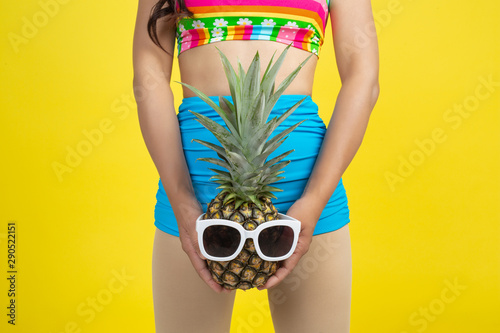 Beautiful woman in a swimsuit holding a pineapple poses on a yellow background.