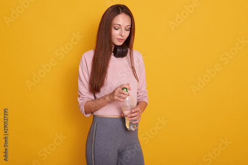 Image of fitness woman standing in studio over yellow background with bottle of water and earphones, looking away, wearing stylish sportwear, having long dark staright hair. Healthy lifesatyle concept photo