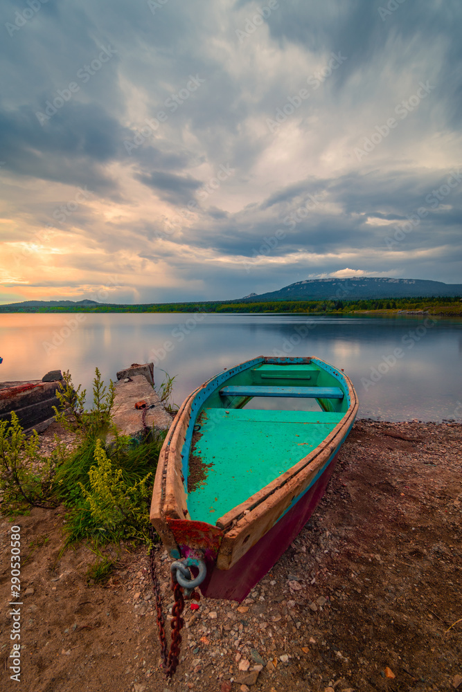 Green wooden boat moored on the shore of lake under cloudy sky at sunset Green wooden boat moored on the shore of lake under cloudy sky at sunset