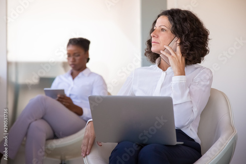 Serious excited businesswoman talking on phone at computer. Business woman sitting in armchair, using laptop and calling on cellphone in office lobby. Communication concept