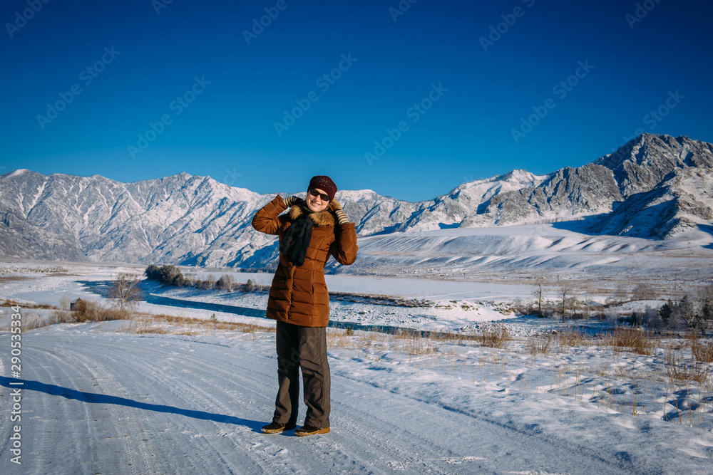 Young cheerful woman in sunglasses stands against snow-covered mountains and blue sky in winter sunny day. Happy girl laughs and looks at camera. Concept of freedom of travel.