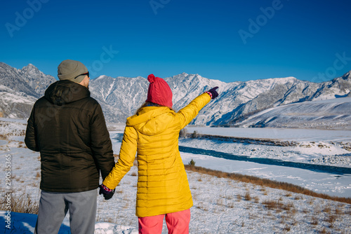 Young happy couple in snowy mountains with copy space. Guy and girl in bright winter clothes look at the snowy peaks. Christmas holidays.