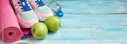Pink yoga mat, sport shoes, bottle of water and apples on blue wooden background. Sport, healthy lifestyle, yoga concept. Female sport equipment. Copy space