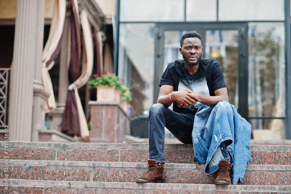 Young attractive serious african american man model. Modern handsome guy in jeans jacket sitting on urban city street. Young attractive serious african american man model. Modern handsome guy in jeans jacket sitting on urban city street.