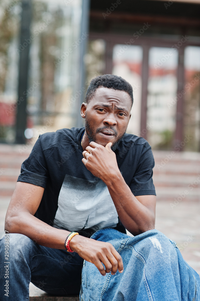 Young attractive serious african american man model. Modern handsome guy in jeans jacket sitting on urban city street. Young attractive serious african american man model. Modern handsome guy in jeans jacket sitting on urban city street.