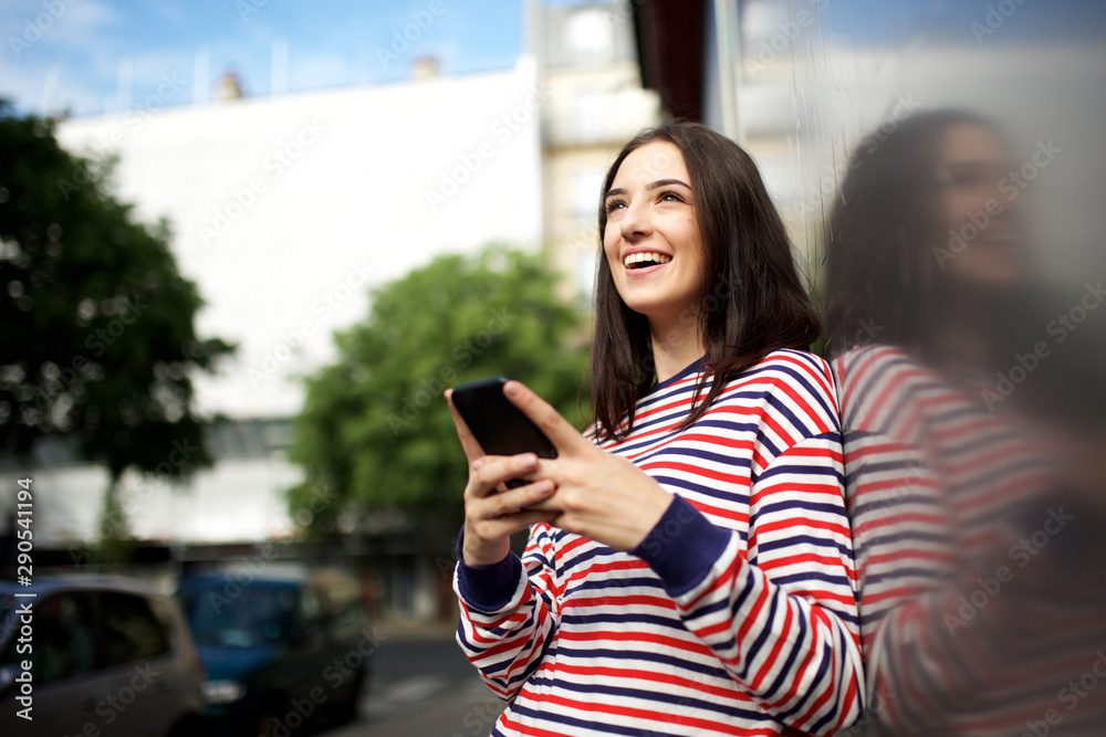 happy young woman with mobile phone leaning against wall outside