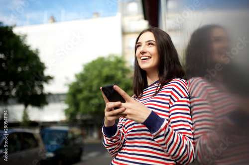happy young woman with mobile phone leaning against wall outside