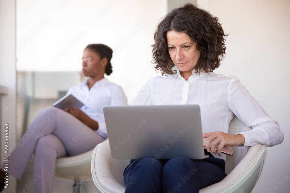 Serious tired businesswoman working on laptop in office lounge. Diverse business women sitting in armchairs, using gadgets in office lobby. Wireless internet connection concept