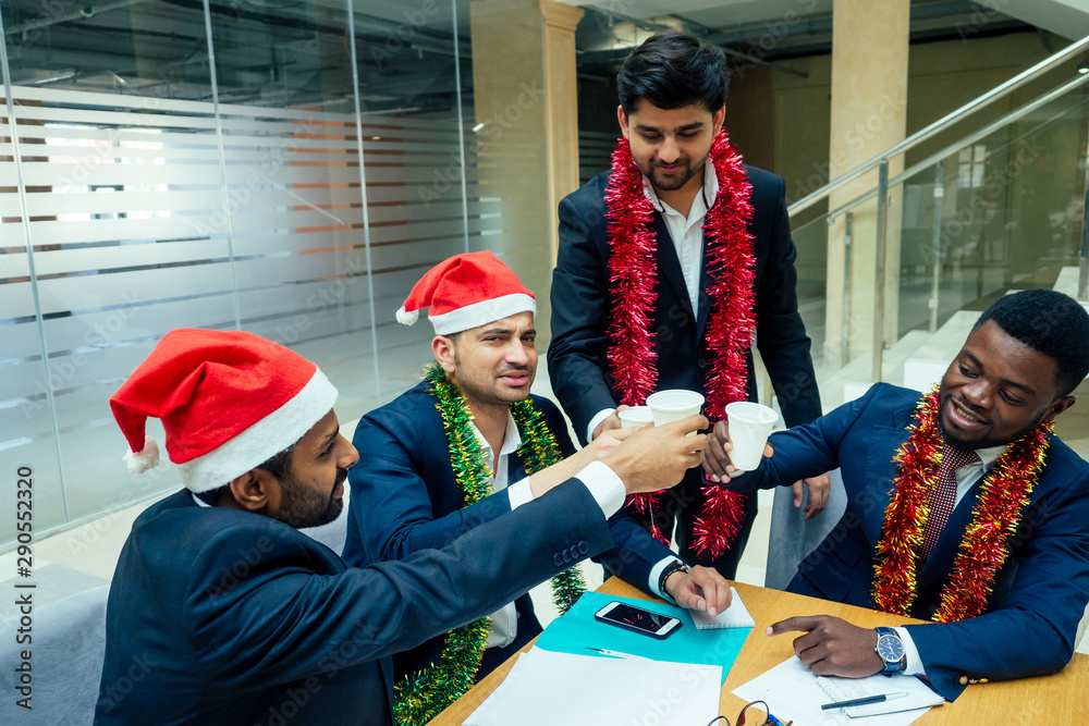 happy group of indian and african american people drinking toast from cup,wearing santa hat and tinsel
