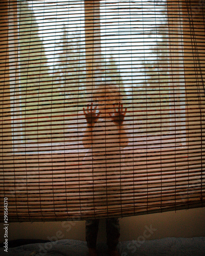 A young person standing behind a bamboo blind with both hands on it.