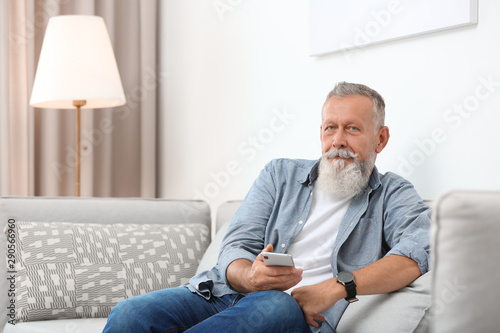 Portrait of handsome mature man with smartphone sitting on sofa in room