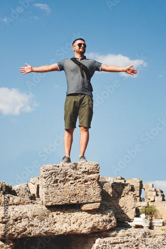 Tourist on the background of the sky with his hands apart. A young man in shorts is facing the camera. Sunny day, travel, joy and achievement.