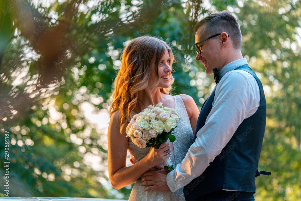 Newly wed couple standing closely together and hugging with smiles. Park with sunset is on background and blurred. Newly wed couple standing closely together and hugging with smiles. Park with sunset is on background and blurred.