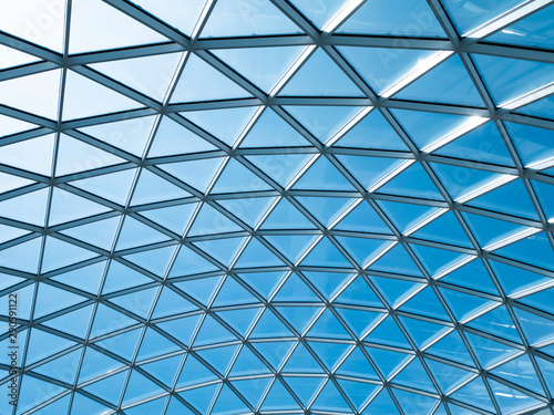 Glass roof with triangular cells and frames against the blue sky with white clouds. Reflections in the glass.