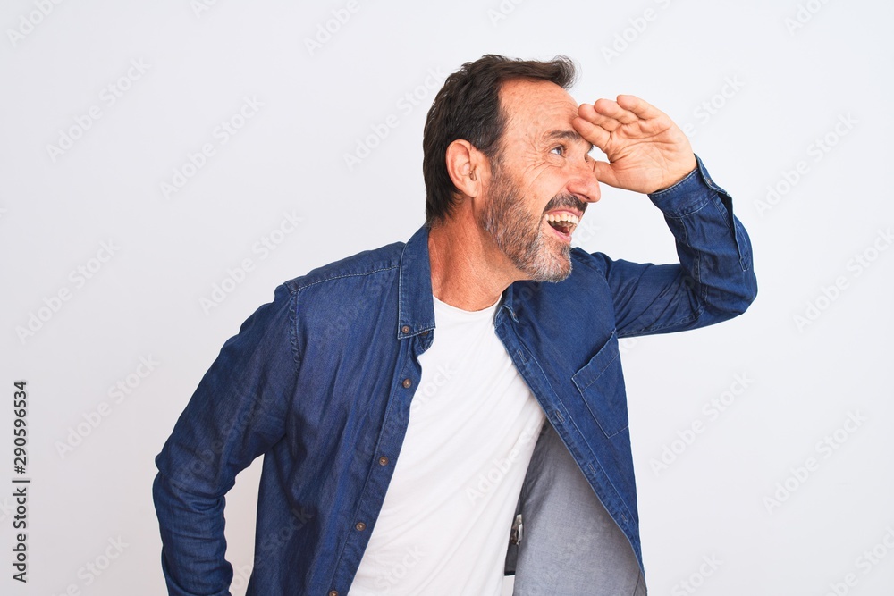 Middle age handsome man wearing blue denim shirt standing over isolated white background very happy and smiling looking far away with hand over head. Searching concept.