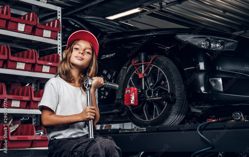 Fototapeta premium Pretty girl is posing for photographer while sitting at dark auto service holding big wrench.
