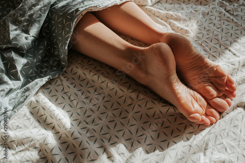 Feet of woman peeking out from under the covers. Girl alone sleep in bed. Wake up. Female legs. Happy morning in bedroom. Cozy and comfortable. Sunlight on bed linen. Pillow, blanket. Recovery, relax