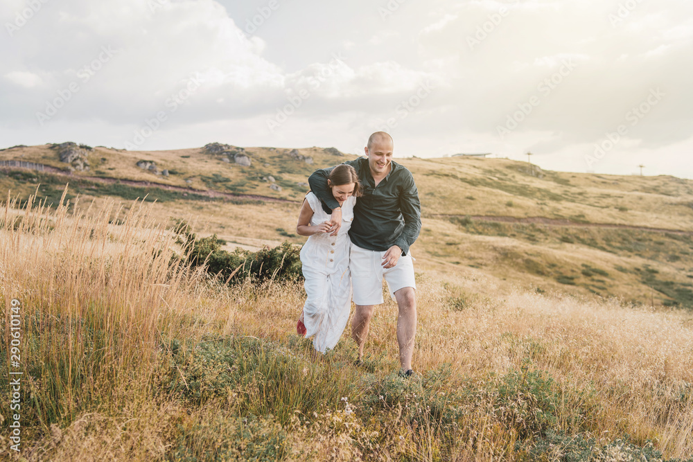 Young couple man and woman in love walking on the field on the mountain in autumn or summer day hugging together having fun bonding flirting flirt husband and wife lovers