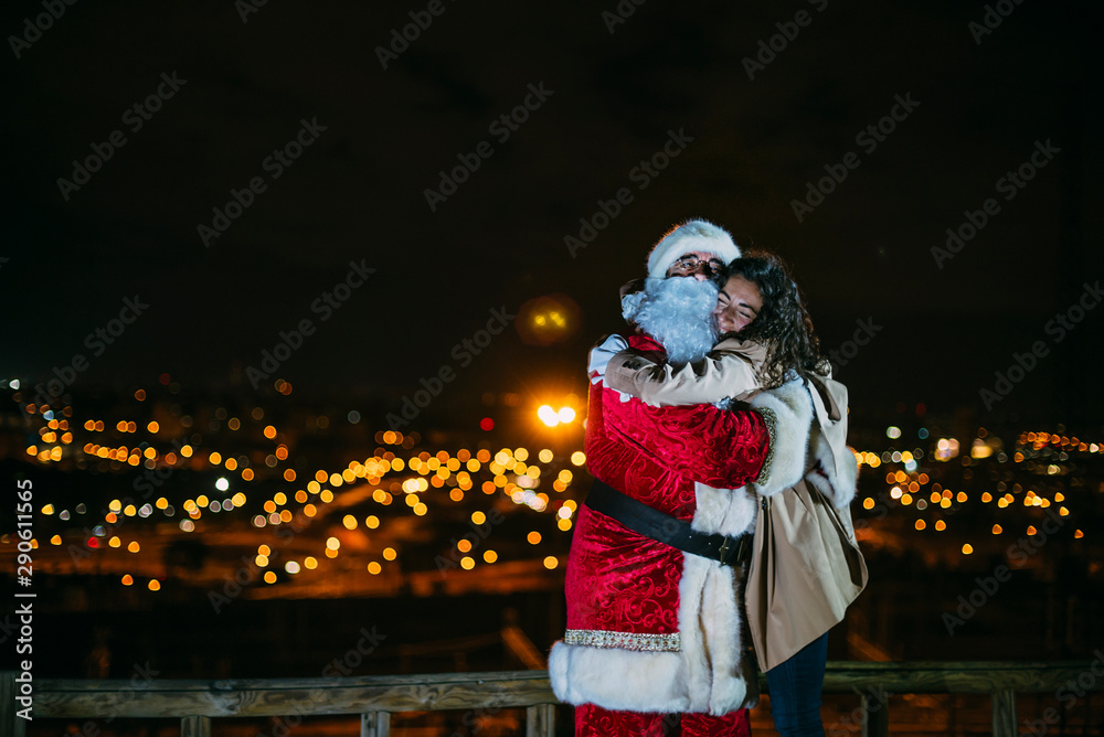 santa claus hugging smiling woman outdoors with lots of christmas lights and christmas presents